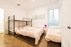 A bedroom with two bunk beds, a single bed, a window with blinds, a pink lamp, and a wall-mounted artwork. - TET (Madrid)
