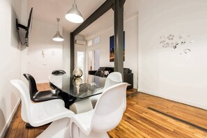 A modern dining area with a glass table and white chairs, a mounted TV, and a decorative wall piece.