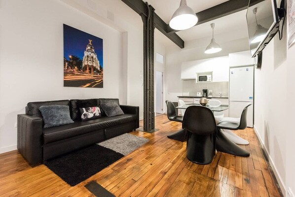 Ein modernes Wohnzimmer mit einer schwarzen Couch, einem Glastisch und einer Küche im Hintergrund.