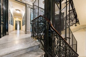 A grand staircase with ornate black metal railings and a wooden handrail, leading to a hallway with classical architecture and a marble floor. - GV1 (Madrid)