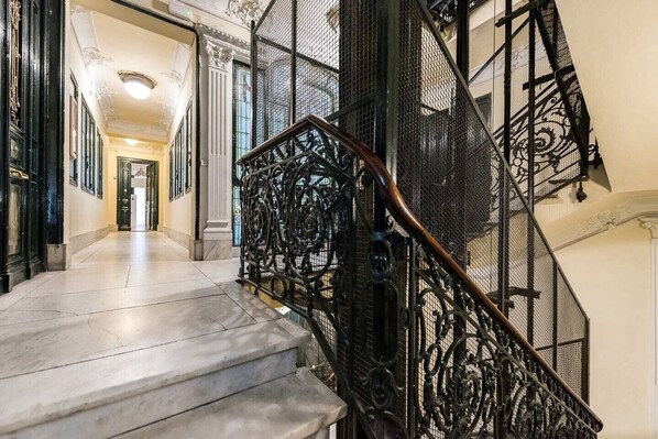 A grand staircase with ornate black metal railings and a wooden handrail, leading to a hallway with classical architecture and a marble floor.