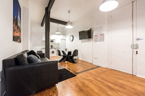 A modern living room with a black sofa, a dining table with chairs, a kitchen area, and a large TV on the wall.