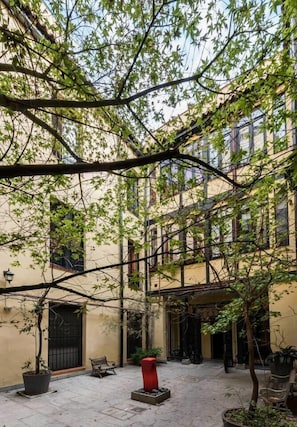 A courtyard with a building, trees, a bench, and a red planter.