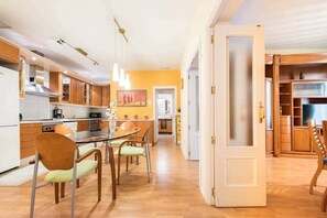A modern kitchen with wooden cabinets, a dining area with chairs, and a hallway leading to a bedroom.