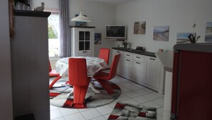 Cocina compacta con armarios blancos, una mesa de comedor con sillas rojas, un televisor y obras de arte en las paredes.