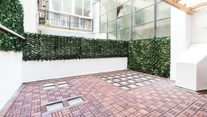 Ein Innenhof mit Pflastersteinen, einer grünen Hecke und einem Fenster mit einem Metallgitter.