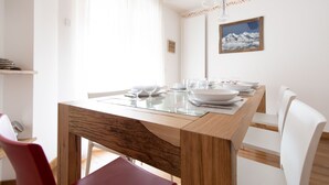 Un comedor con una mesa de madera dispuesta para cuatro personas, sillas blancas y un cuadro de un paisaje montañoso en la pared.