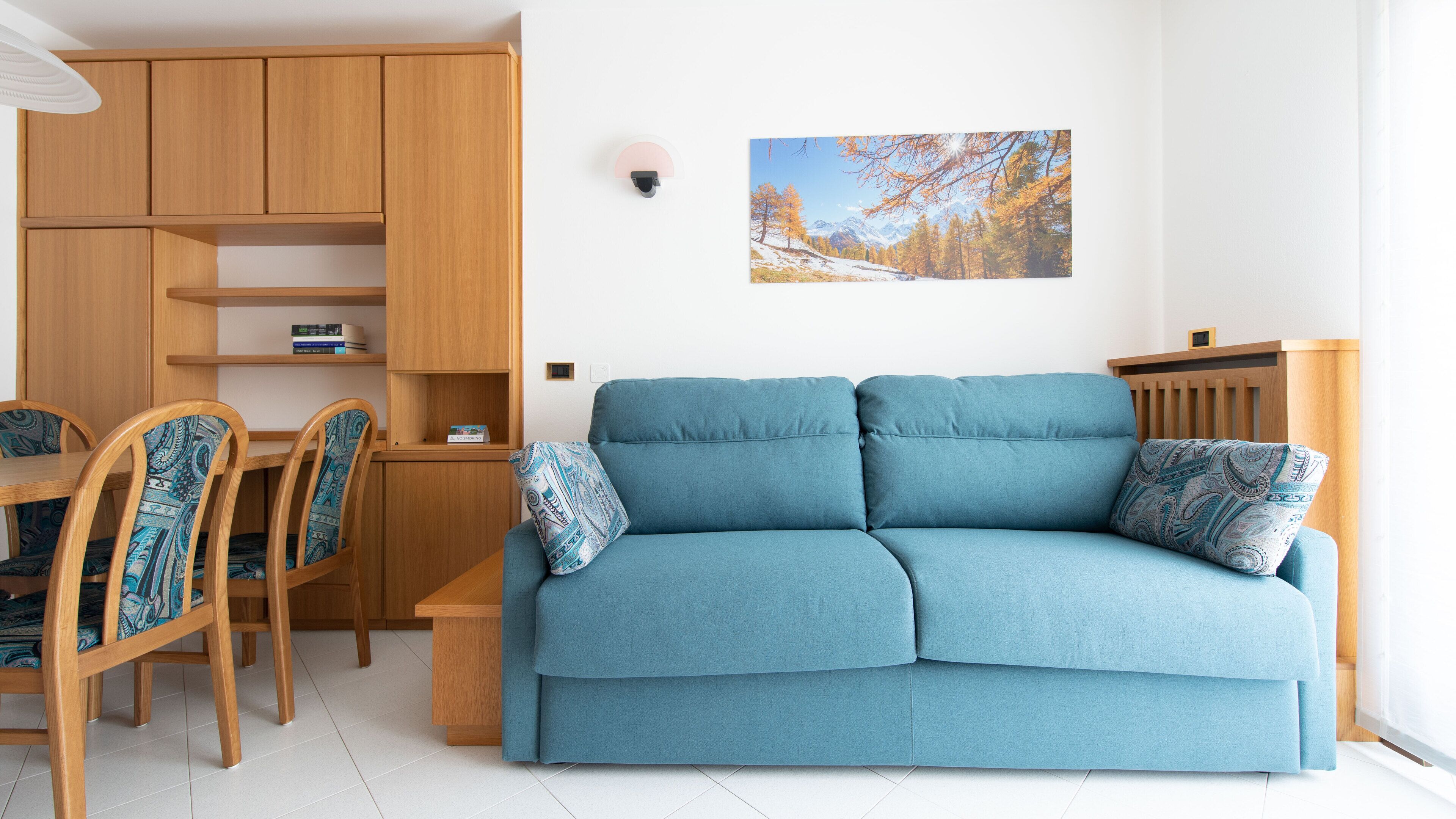A living room with a blue sofa, wooden chairs, a wooden dining table, and a wall-mounted lamp.