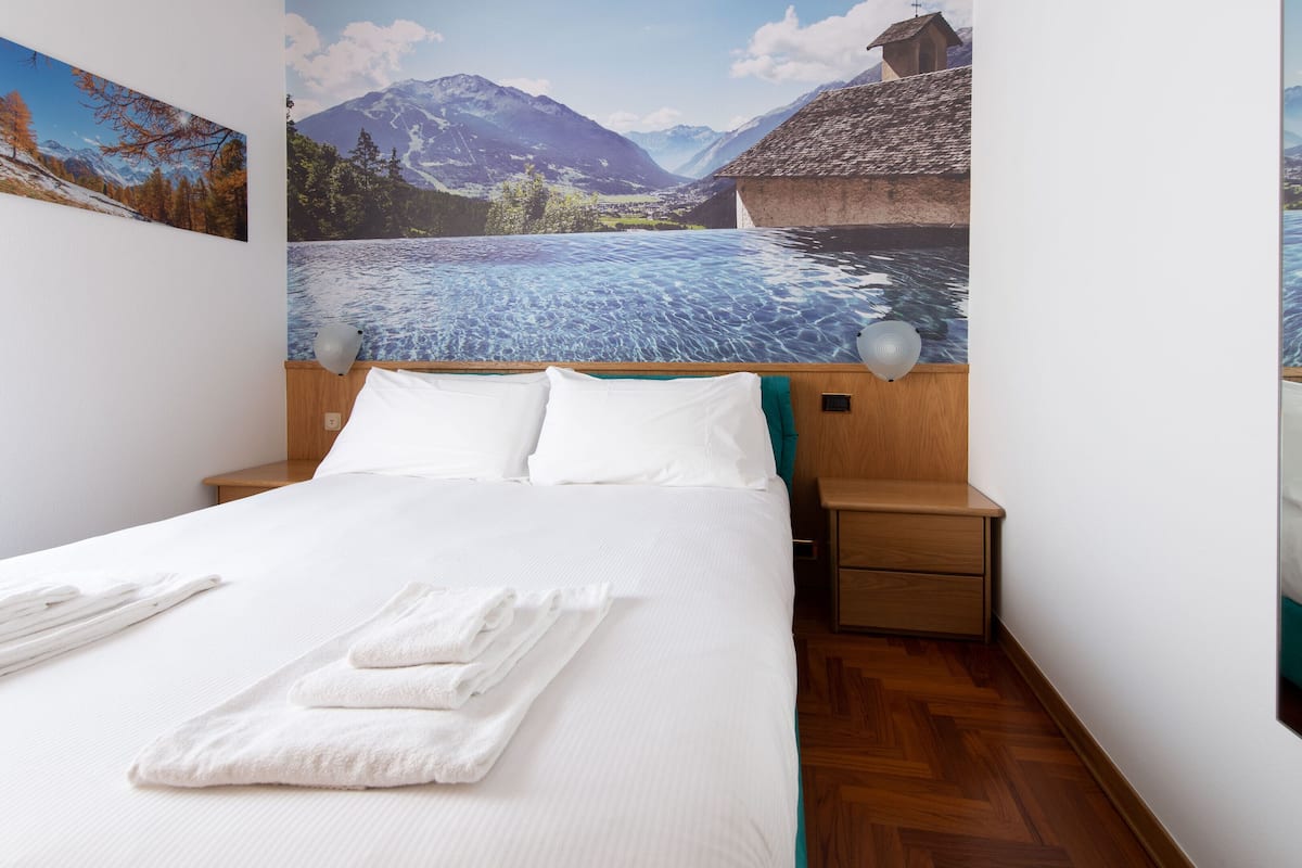 Ein Hotelzimmer mit einem Bett, einem Nachttisch und einer Wandmalerei, die eine Berglandschaft zeigt.