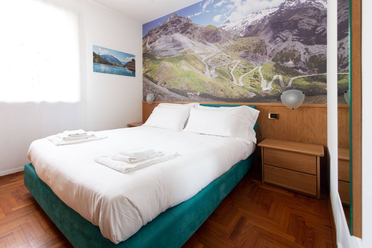 Ein ordentlich bezogenes Bett mit weißen Leinentüchern und einem türkisfarbenen Bettrunden, ein Nachttisch aus Holz mit einer Lampe und ein großes Gemälde mit einer Berglandschaft an der Wand.