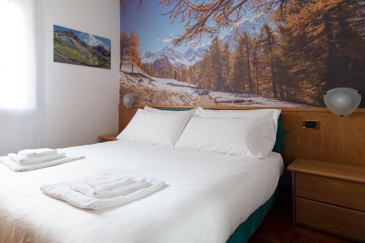 Ein ordentlich bezogenes Bett mit weißen Leinen und Handtüchern, ein hölzerner Kopfseitenverzierer und ein an der Wand befestigter Nachttisch mit einer Lampe.