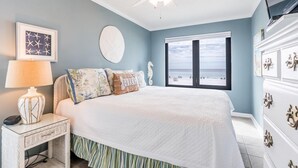 A bedroom with a bed, a ceiling fan, a nightstand with a lamp, a dresser, and a large window with a view of the beach. - Cozy Gulf Front Condo With Beach Front View | Beach Access | Pool | Sleeps 6 (Gulf Shores)