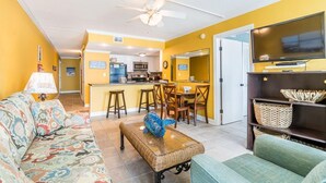 A living room with a sofa, a coffee table, and a television. There is a kitchen in the background with a microwave and a refrigerator. - Cozy Gulf Front Condo With Beach Front View | Beach Access | Pool | Sleeps 6 (Gulf Shores)