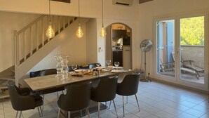 Un espace repas comprenant une longue table, quatre chaises, des luminaires suspendus, un escalier et une vue à travers une porte vitrée.