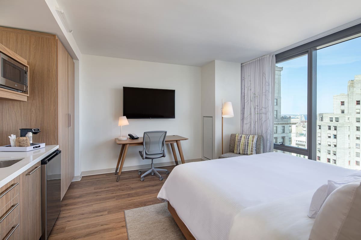 A hotel room with a bed, a desk with a chair, a television, and a large window with a city view.