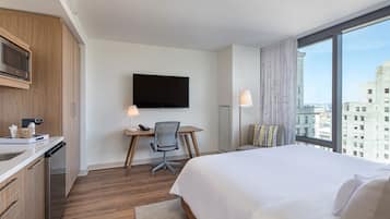 A hotel room with a bed, a desk with a chair, a television, and a large window with a city view.