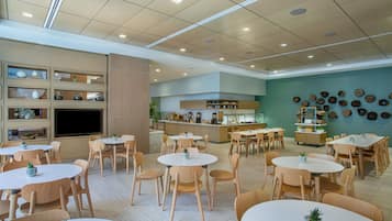 A spacious dining area with multiple round tables and chairs, a kitchen area with a counter, and a wall decorated with various items.