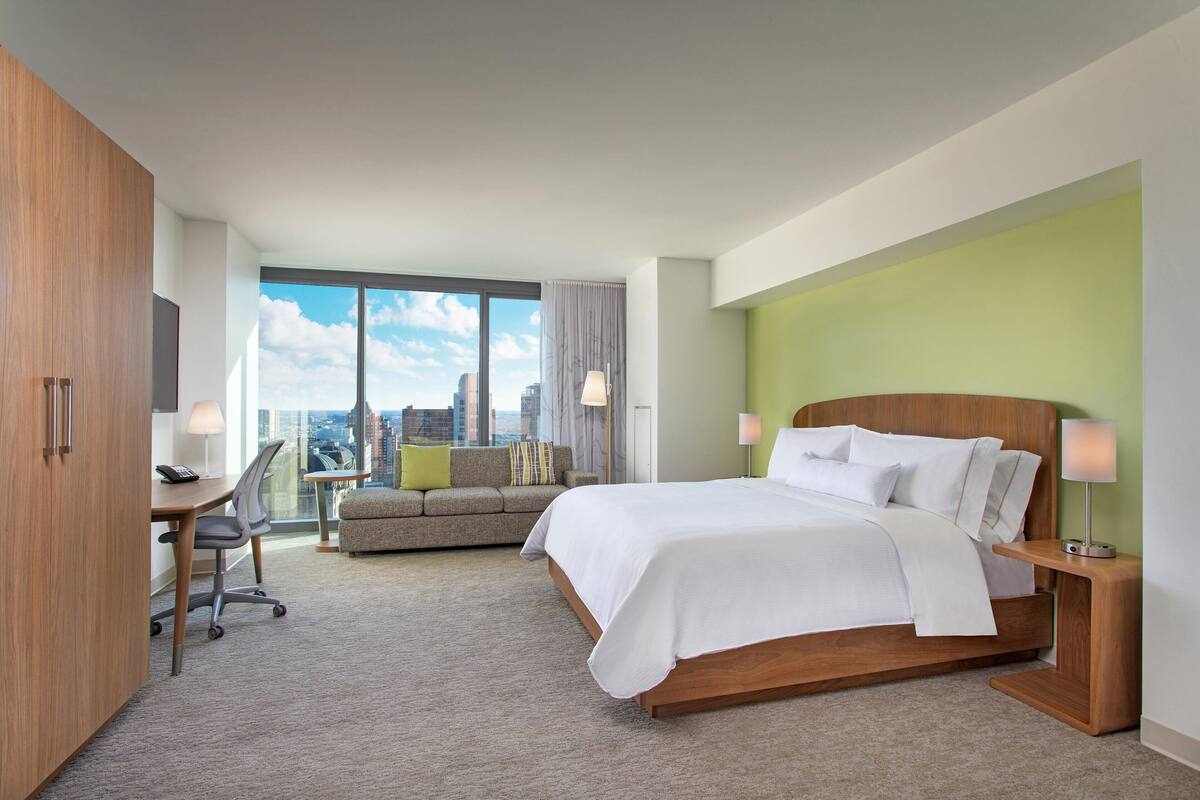 A modern hotel room with a large bed, a desk, and a sofa. The room has a city view through large windows.