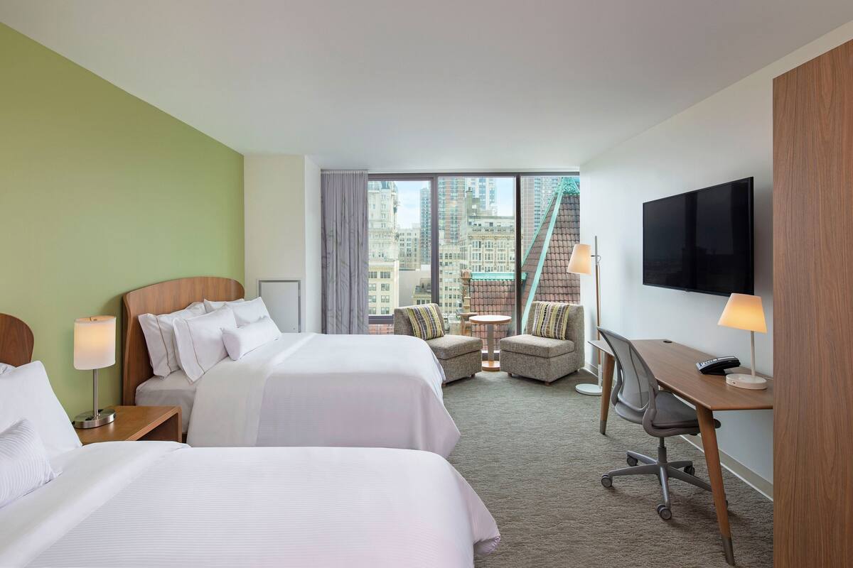 A hotel room with two beds, a desk, a chair, and a TV.