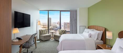 A hotel room with two beds, a desk, a chair, a TV, and a view of the city.