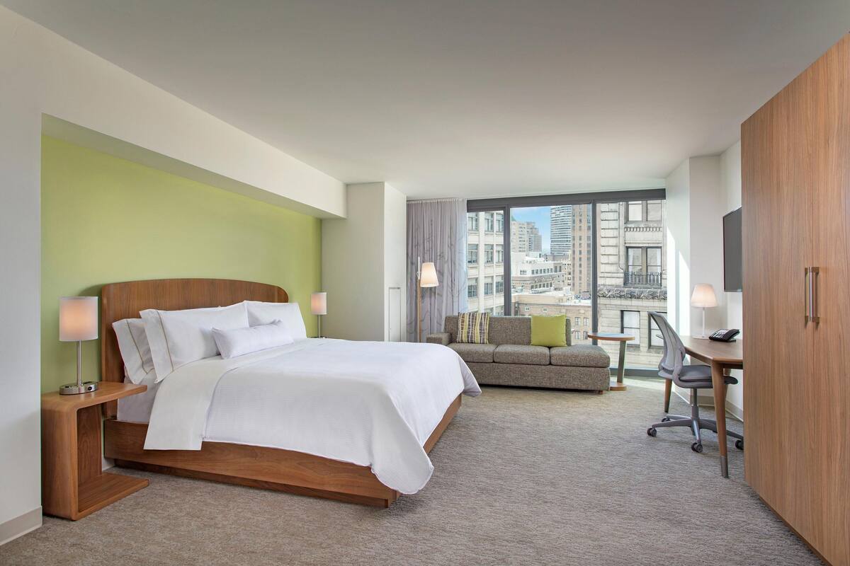A modern hotel room with a large bed, a sofa, a desk, and a view of the city through large windows.