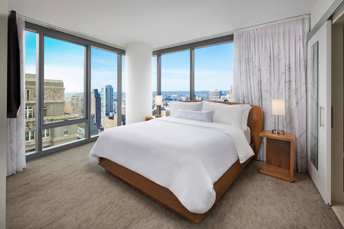 A modern hotel room with a large bed, a nightstand, and a view of the cityscape through floor-to-ceiling windows.