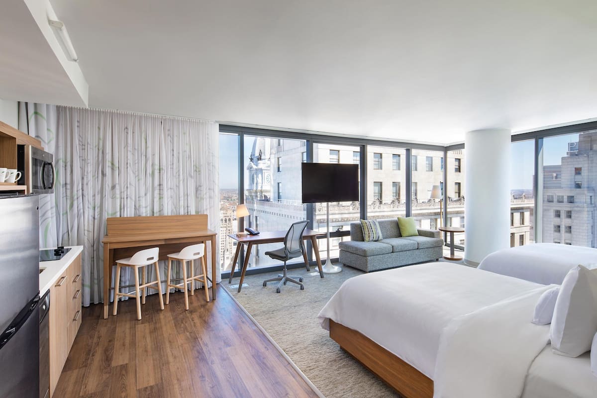 A modern hotel room with a large bed, a desk with two chairs, a sofa, a TV, and a city view.