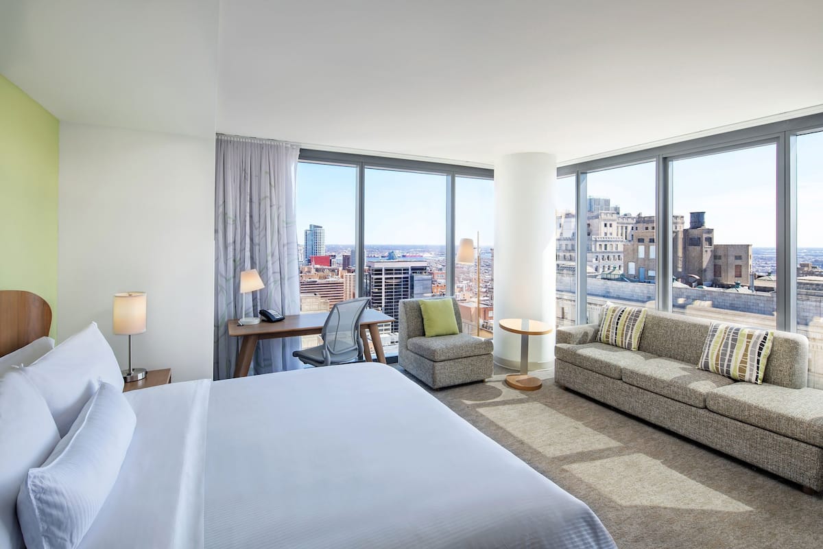 A hotel room with a large bed, a sofa, a desk, and a chair. There is a city view through large windows.