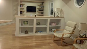 A living room with a wooden floor, a white armchair, a small table with a lamp, and a TV on a shelf.