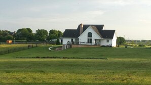 A large white house with a prominent front porch and a large window, surrounded by a fenced property and open fields.