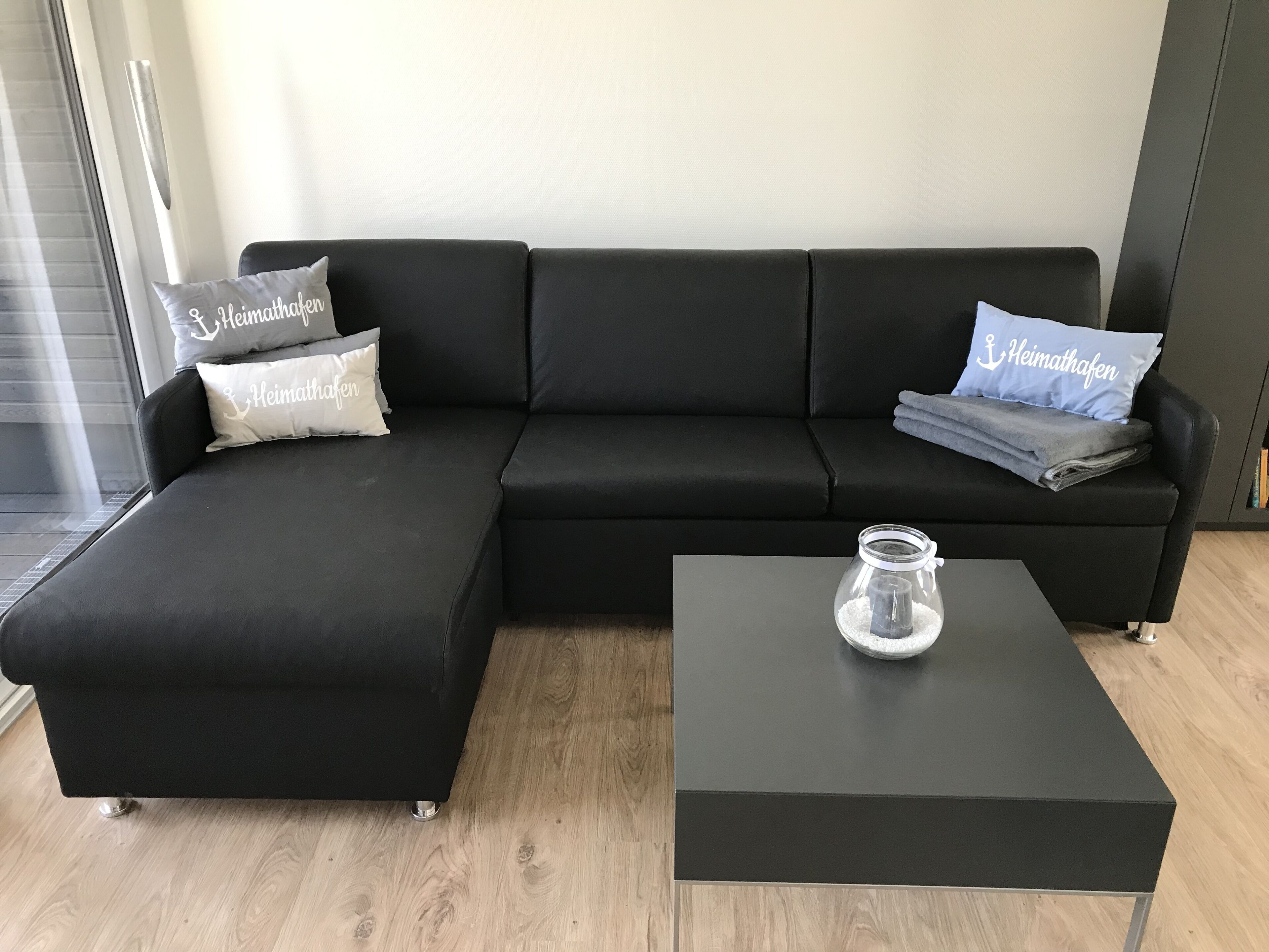 A modern living room with a black sectional sofa, two pillows with "Heimathafen" text, a glass jar on a black coffee table, and a wooden floor.