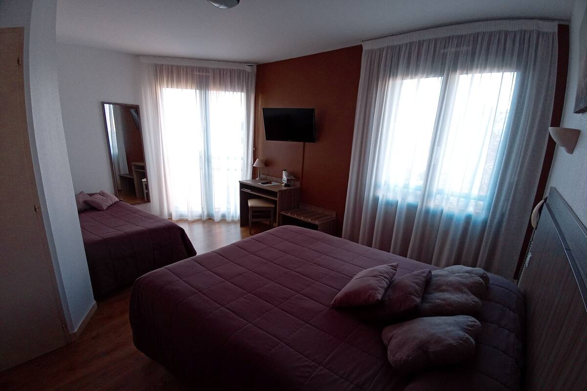 Habitación de hotel con dos camas, un escritorio, una silla, un televisor y ventanas amplias con cortinas.
