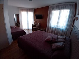 Une chambre d’hôtel avec deux lits, un bureau, une chaise, une télévision et de grandes fenêtres avec des rideaux.