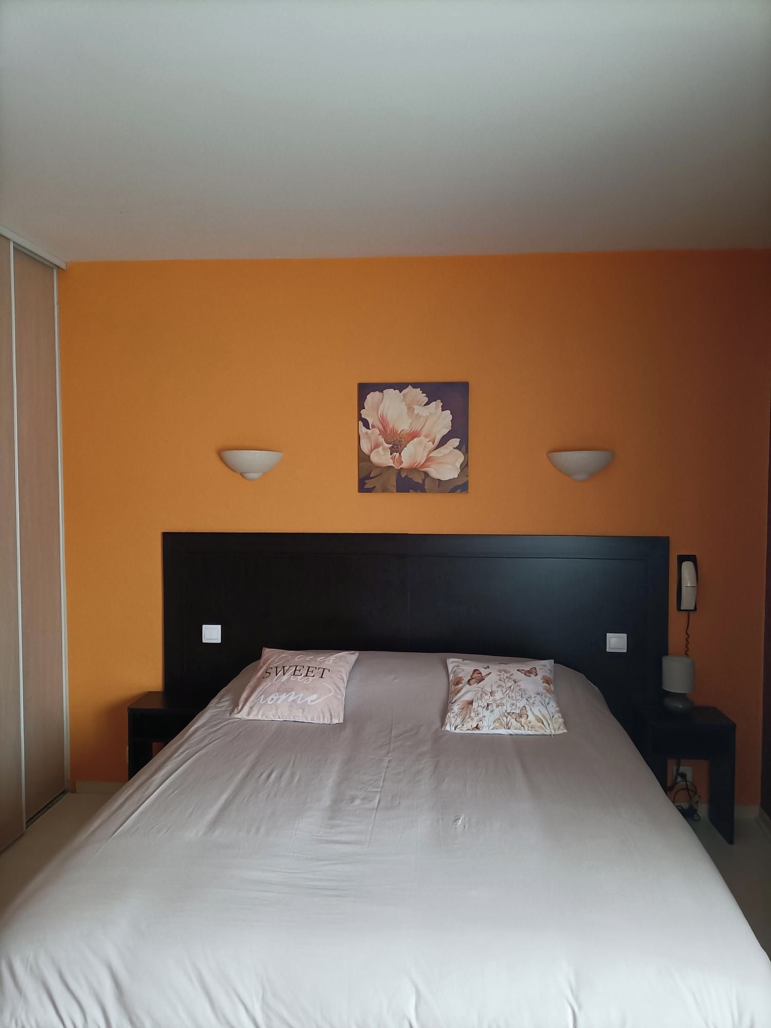 A bedroom with a large bed, two bedside tables, a nightstand, a wall-mounted telephone, and a floral painting above the bed.