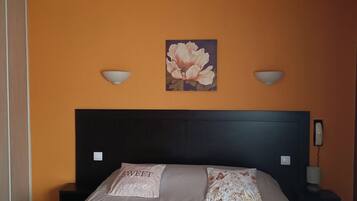 A bedroom with a large bed, two bedside tables, a nightstand, a wall-mounted telephone, and a floral painting above the bed.