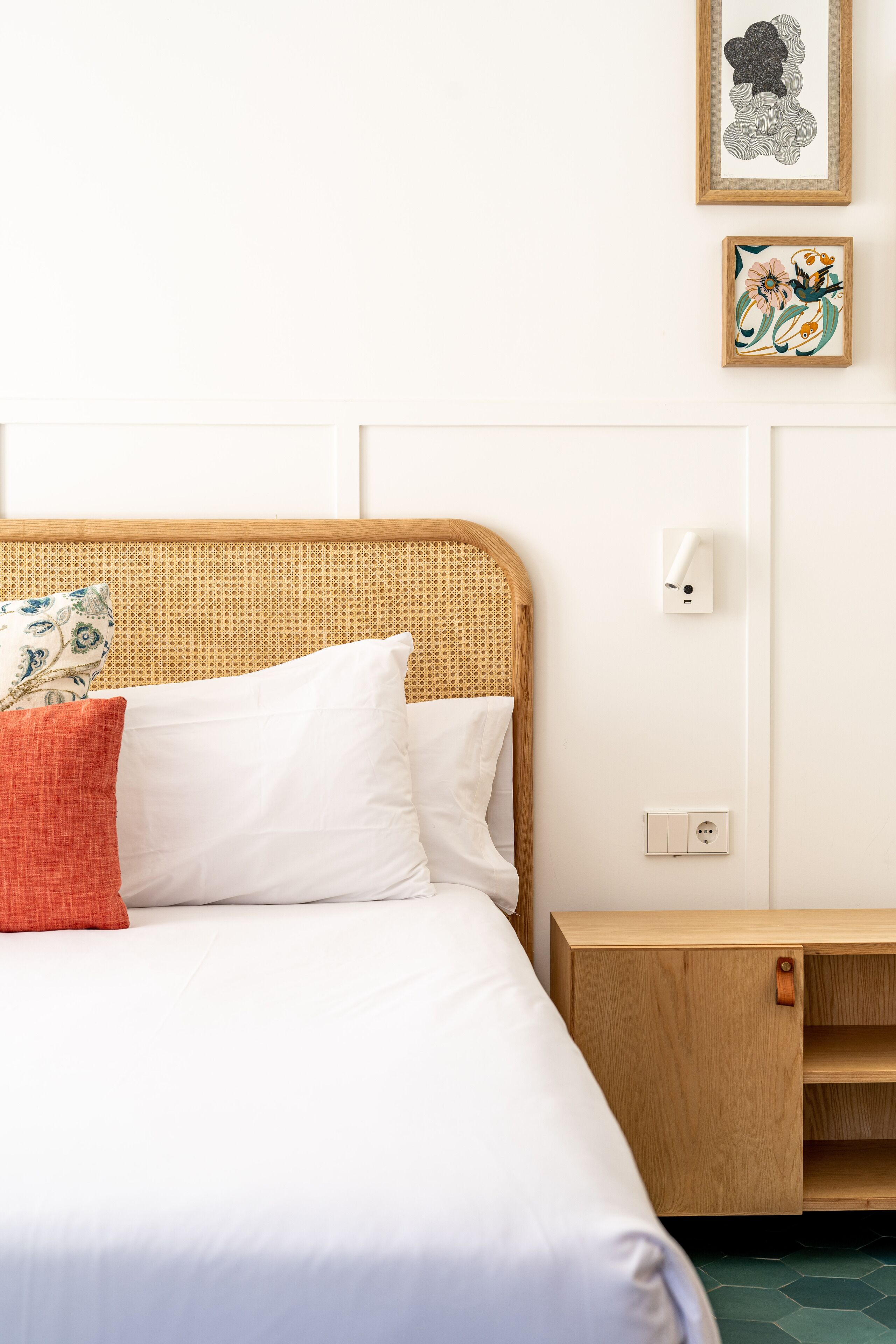 Una cama bien tendida con sábanas blancas, cabecera de madera y almohada roja. Una pequeña mesita de noche con cajón se encuentra al lado de la cama.