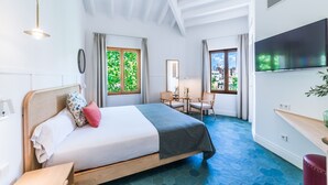 Habitación de hotel con cama, escritorio, silla, televisión y ventanas grandes con cortinas.