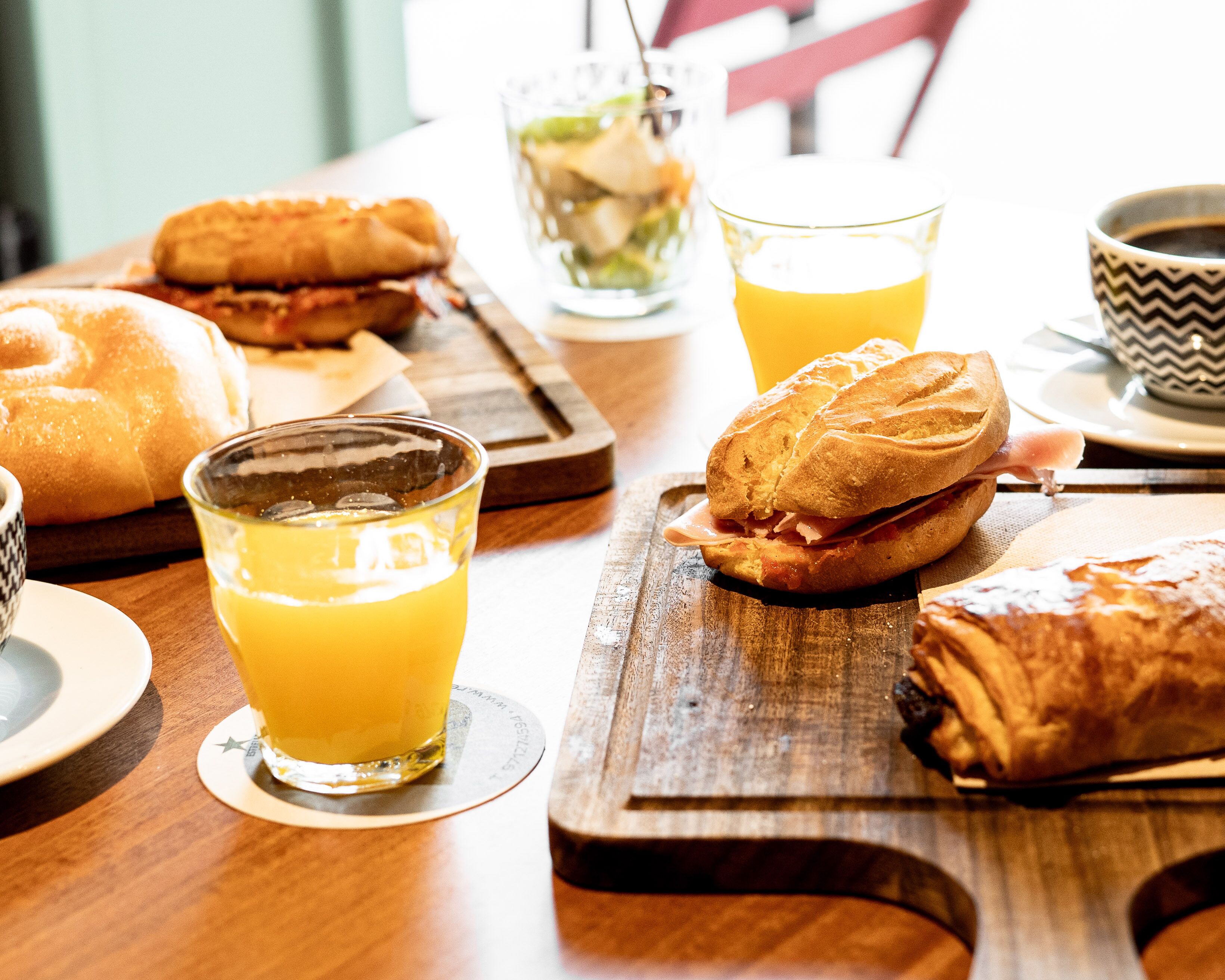 Une table en bois dressée pour le petit-déjeuner, avec des sandwichs, des croissants et du jus d’orange.