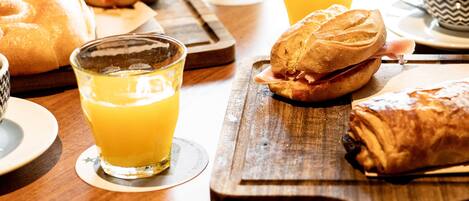 Une table en bois dressée pour le petit-déjeuner, avec des sandwichs, des croissants et du jus d’orange.