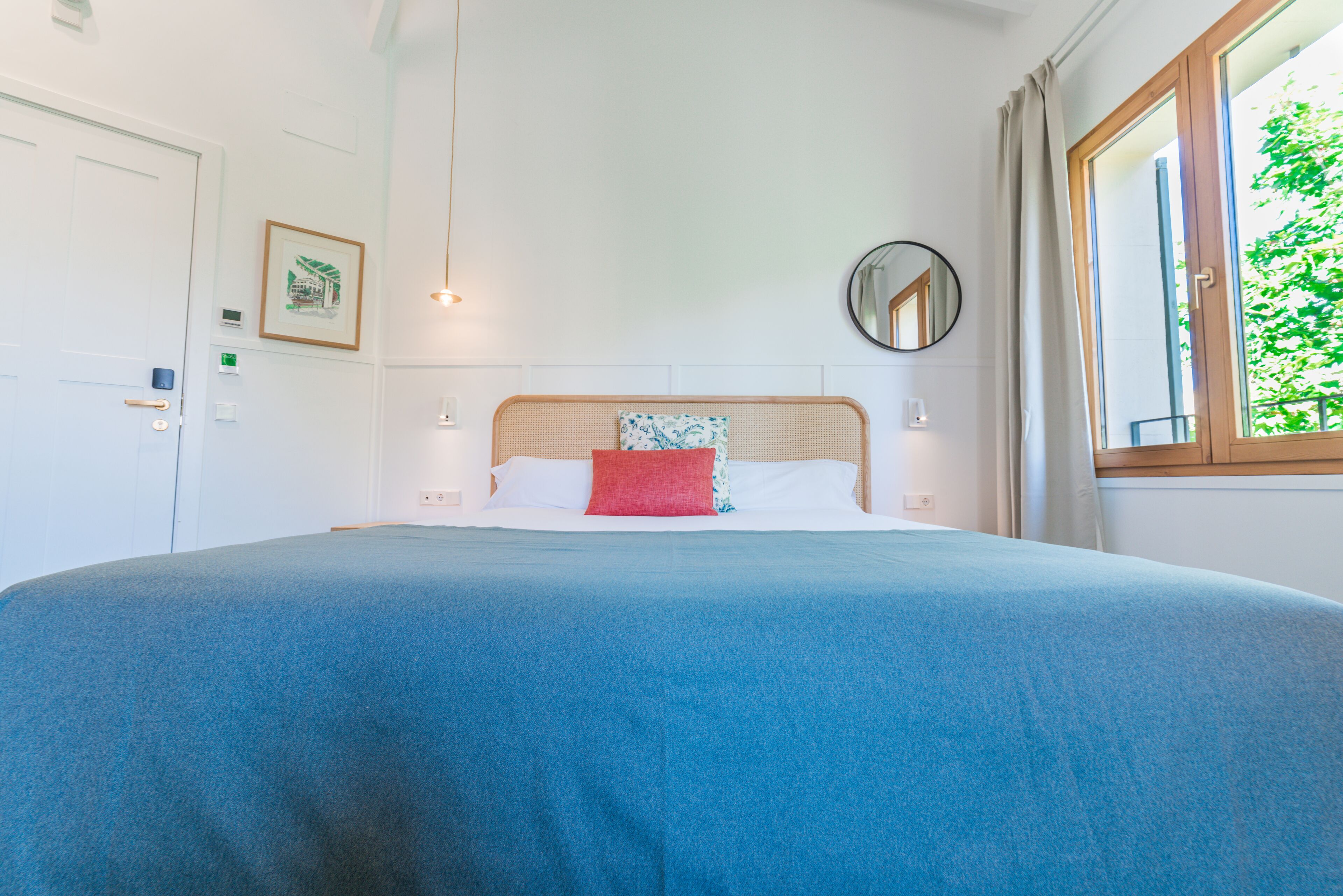 A neatly made bed with a blue bedspread and a red pillow, a round mirror on the wall, and a window with curtains.
