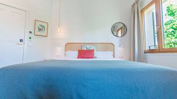 A neatly made bed with a blue bedspread and a red pillow, a round mirror on the wall, and a window with curtains.