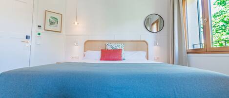 A neatly made bed with a blue bedspread and a red pillow, a round mirror on the wall, and a window with curtains.