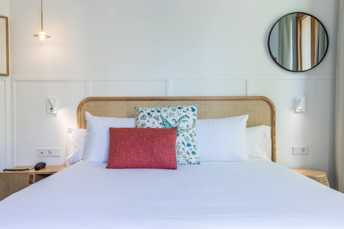 A neatly made bed with white linens and a red pillow, a wicker headboard, a round mirror on the wall, and a bedside table with a phone and a notepad.