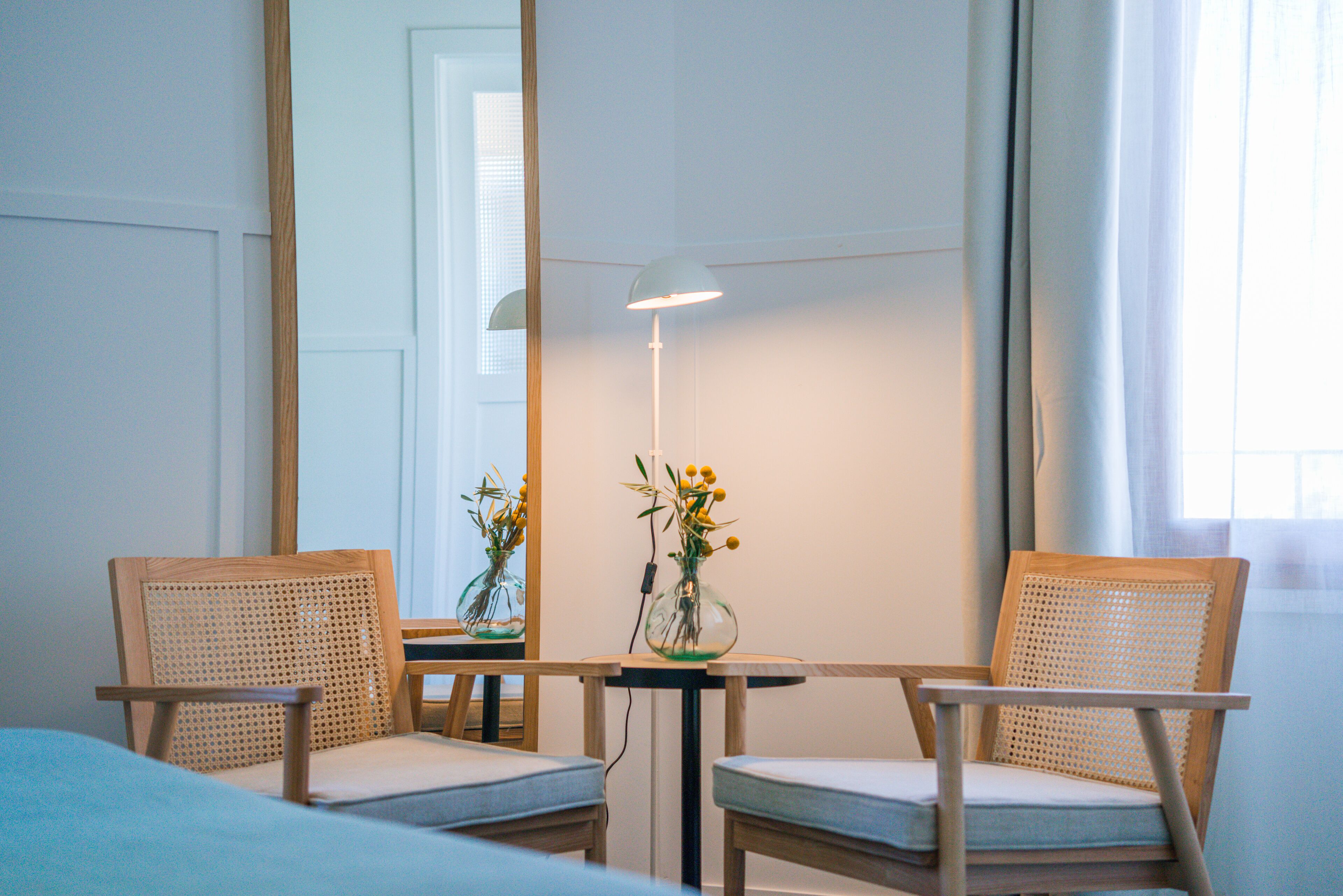 A hotel room with two wooden chairs, a small table, a lamp, and a vase with flowers.