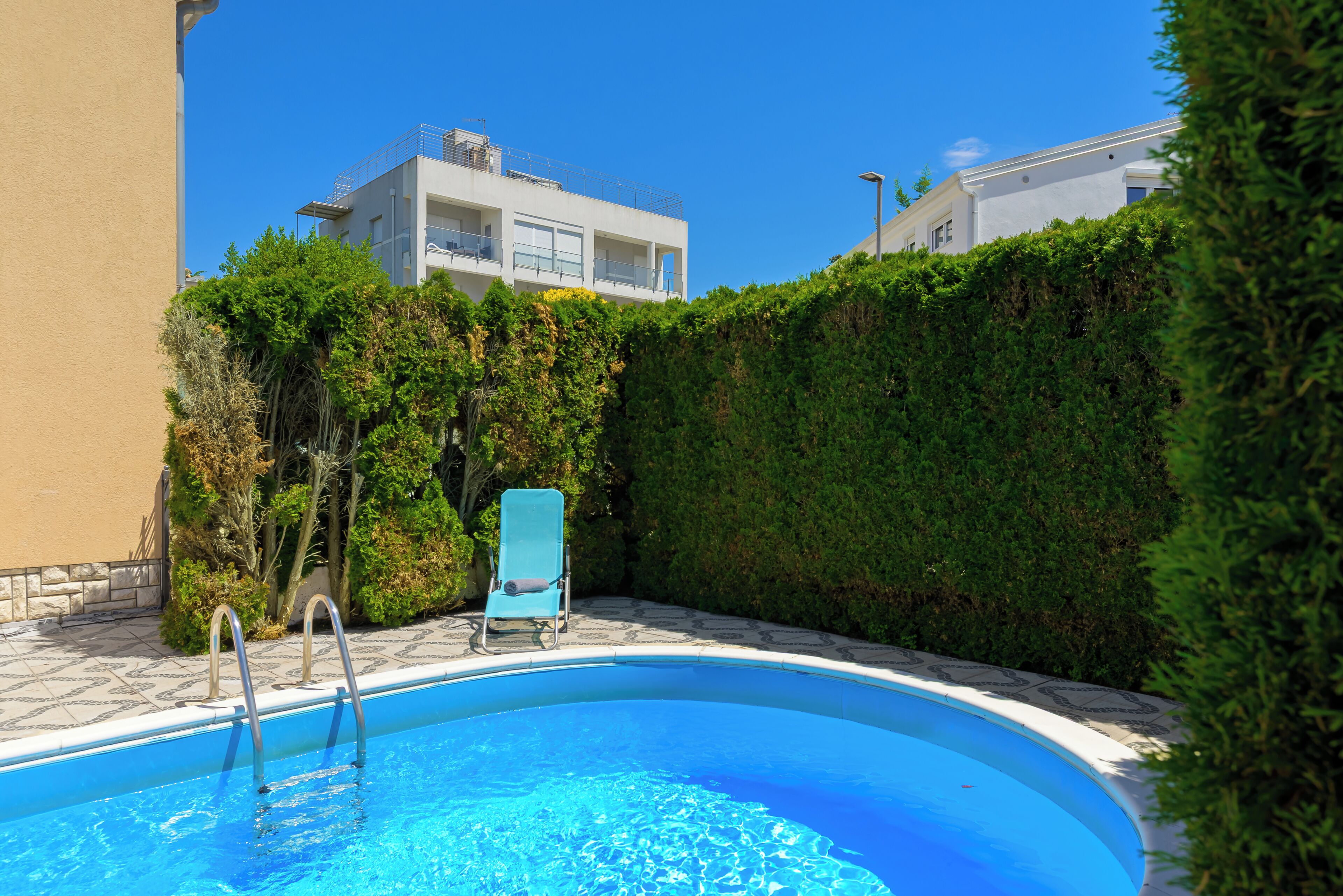 A swimming pool with a blue water feature, surrounded by a hedge and a building with balconies.