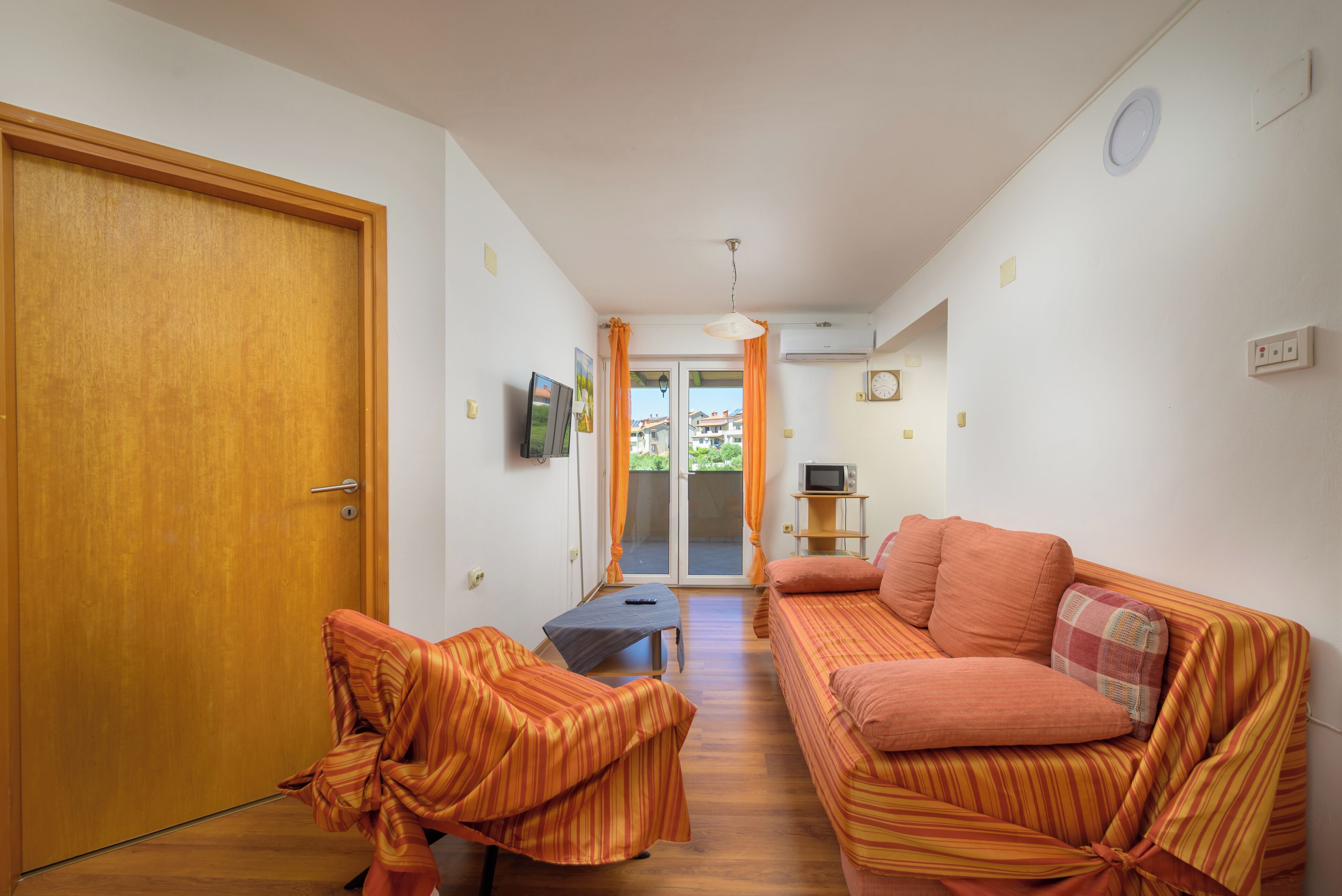 A room with a wooden door, a sofa, a television, a small table, and a window with curtains.