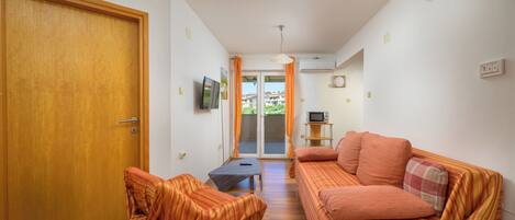 A room with a wooden door, a sofa, a television, a small table, and a window with curtains.