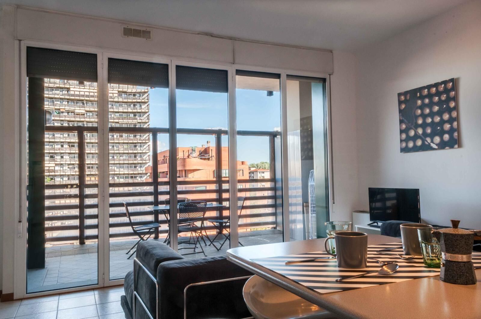 A modern living room with a large sliding glass door leading to a balcony, a dining table with chairs, and a flat-screen TV.
