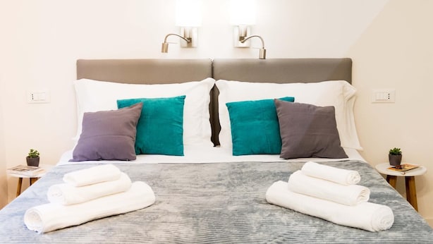 Un lit bien fait, avec une tête de lit grise, des oreillers blancs et turquoise, et deux petites tables de chevet ornées de plantes en pot.