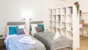 Une chambre d’hôtel avec deux lits, chacun recouvert d’une couverture et doté d’oreillers gris, une étagère blanche avec des livres et une plante, et une petite table sur laquelle est posée une plante.